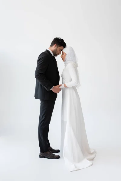 Vista lateral de la pareja musulmana casada cogida de la mano y de pie sobre gris - foto de stock