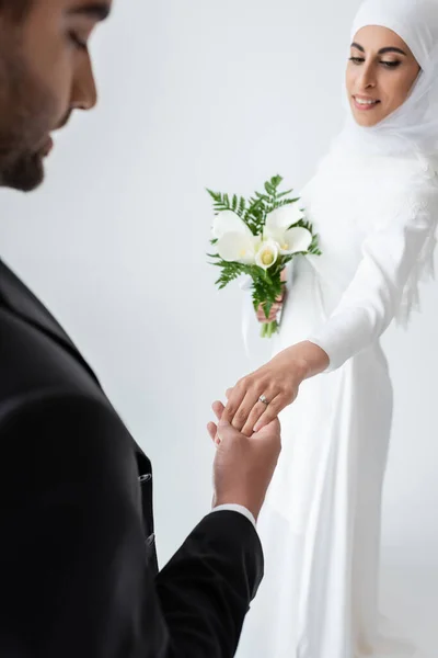 Happy muslim bride with bouquet holding hands with blurred groom isolated on grey — Stock Photo