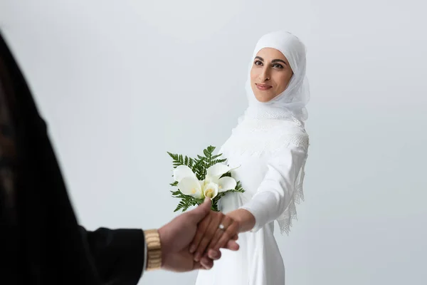 Счастливая мусульманская невеста с букетом, держась за руки с женихом, изолированным на сером — стоковое фото
