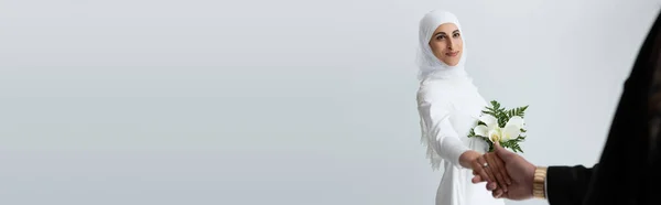 Glückliche muslimische Braut mit Strauß Händchen haltend mit verschwommenem Bräutigam isoliert auf grau, Banner — Stockfoto