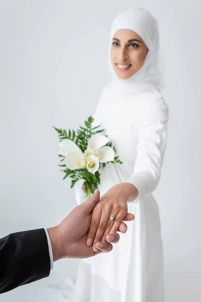 Fröhliche muslimische Braut mit Strauß Händchen haltend mit verschwommenem Bräutigam isoliert auf grau — Stockfoto