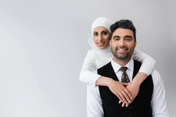 Happy muslim bride in hijab hugging cheerful groom in suit isolated on grey — Stock Photo