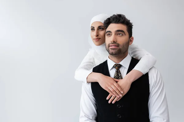 Lächelnde muslimische Braut im Hijab umarmt fröhlichen Bräutigam im Anzug isoliert auf grau — Stockfoto