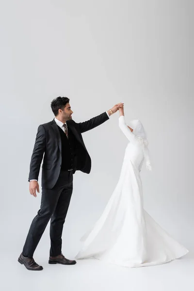 Longitud completa del novio musulmán en traje y novia en vestido blanco bailando en gris - foto de stock