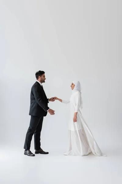 Full length of muslim groom in suit and bride in wedding dress dancing on grey — Stock Photo