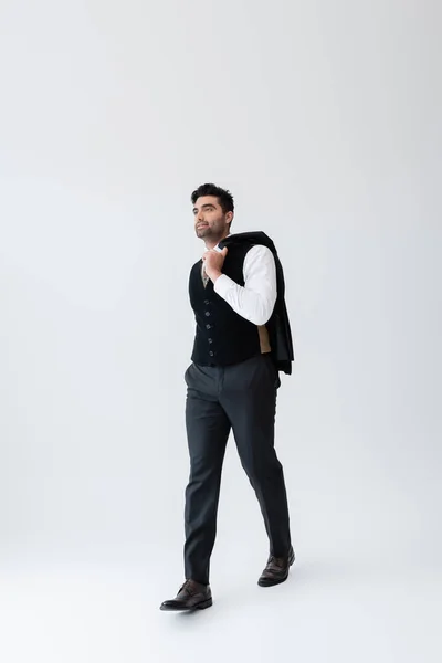 Full length of muslim man in vest walking while holding blazer on grey — Stock Photo