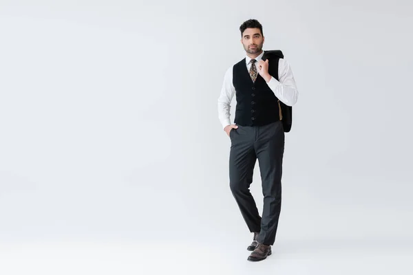 Longitud completa del hombre musulmán en chaleco de pie con la mano en el bolsillo y la celebración de chaqueta aislado en gris - foto de stock