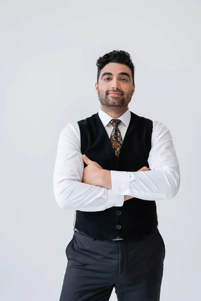 Homme musulman heureux en gilet debout avec les bras croisés isolés sur gris — Photo de stock