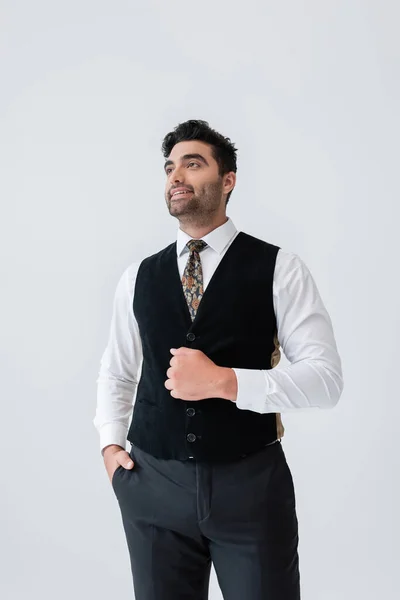 Happy muslim man in vest standing with hand in pocket and smiling isolated on grey — Stock Photo