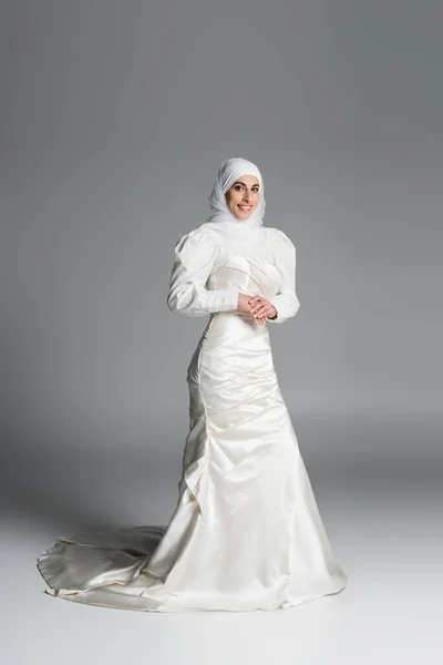 Full length of happy muslim bride in wedding dress and hijab standing on dark grey — Stock Photo