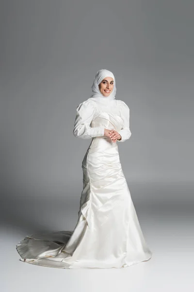 Pleine longueur de mariée musulmane heureuse en robe de mariée et hijab debout sur gris foncé — Photo de stock