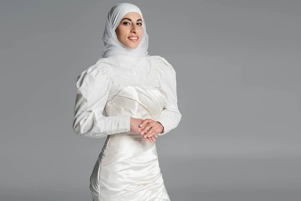 Feliz novia musulmana en vestido de novia y hijab sonriendo en gris - foto de stock