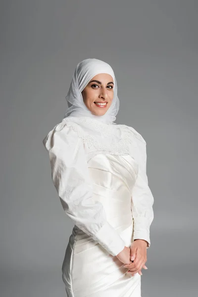 Novia musulmana positiva en vestido de novia y hijab sonriendo en gris oscuro - foto de stock