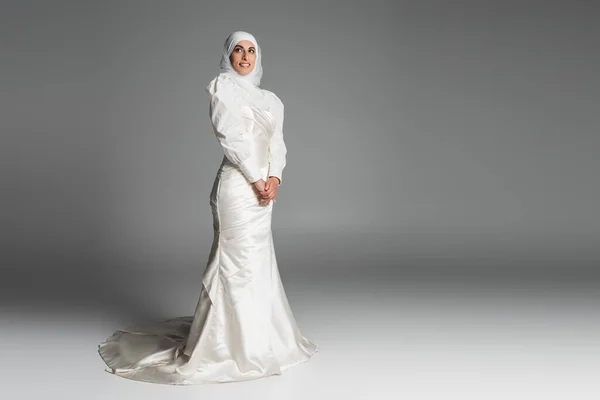 Pleine longueur de mariée musulmane heureuse en robe de mariée et hijab debout sur gris foncé — Photo de stock