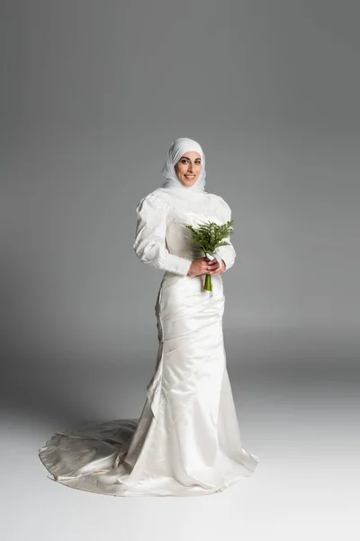 Pleine longueur de mariée musulmane heureuse en robe blanche et hijab tenant bouquet de mariage sur gris foncé — Photo de stock