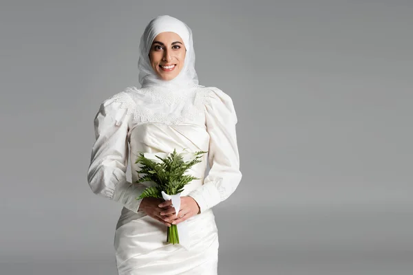 Joyful muslim bride in white dress and hijab holding wedding bouquet on dark grey — Stock Photo