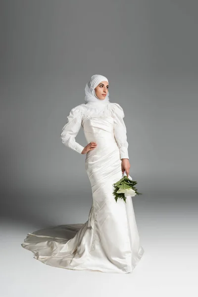 Full length of muslim bride in white dress and hijab holding wedding bouquet while posing with hand on hip on dark grey — Stock Photo