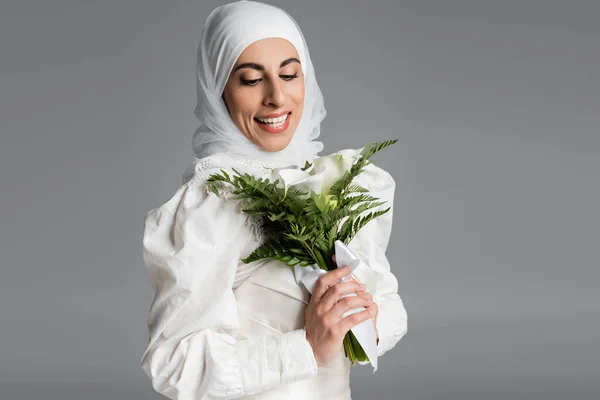 Noiva muçulmana feliz em vestido branco e hijab olhando para o buquê de casamento em cinza escuro — Fotografia de Stock