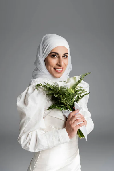 Femme musulmane heureuse en robe blanche et hijab tenant bouquet de mariage sur gris foncé — Photo de stock