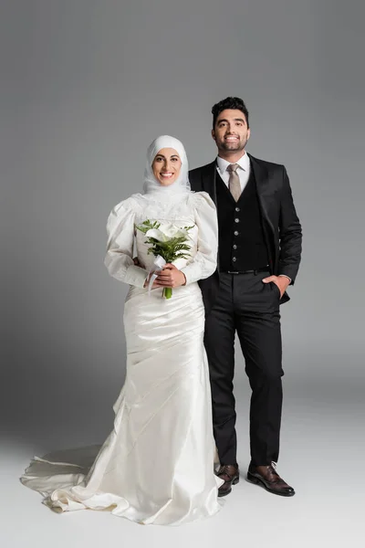 Fröhlicher Bräutigam im Anzug posiert neben muslimischer Braut mit Brautstrauß auf grau — Stockfoto