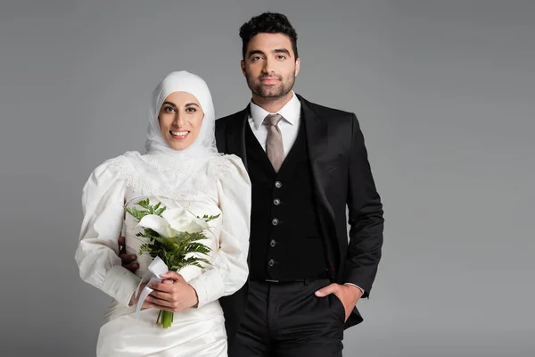 Счастливый жених в костюме позирует рядом с мусульманской невестой со свадебным букетом, изолированным на сером — стоковое фото