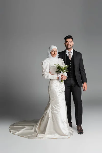 Longitud completa de novio feliz en traje cerca de la novia musulmana con ramo de boda en gris - foto de stock