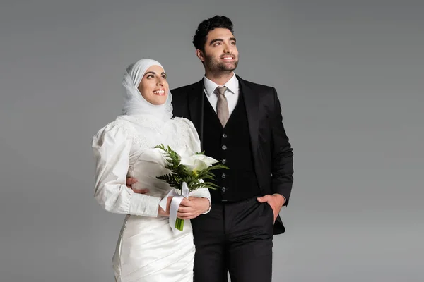 Жених в костюме позирует рядом с мусульманской счастливой невестой со свадебным букетом на сером — стоковое фото