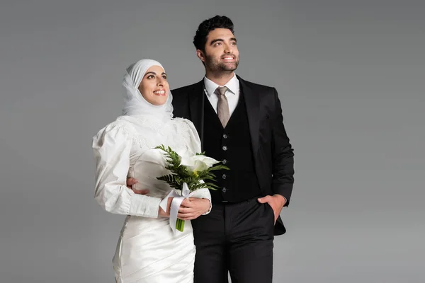 Groom in suit posing near muslim happy bride with wedding bouquet on grey — Stock Photo