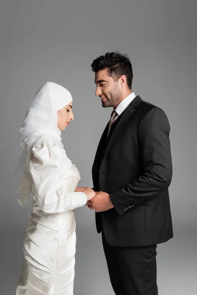 Мусульманская невеста в свадебном платье и веселый жених в костюме держась за руки изолированные на сером — стоковое фото