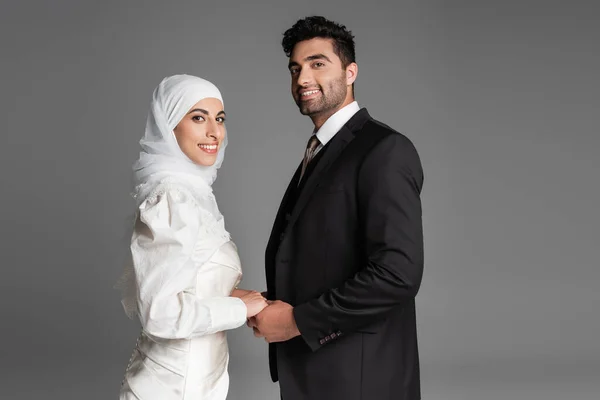 Glückliche muslimische Braut im Hochzeitskleid und Bräutigam im Anzug, die Hände isoliert auf grau — Stockfoto