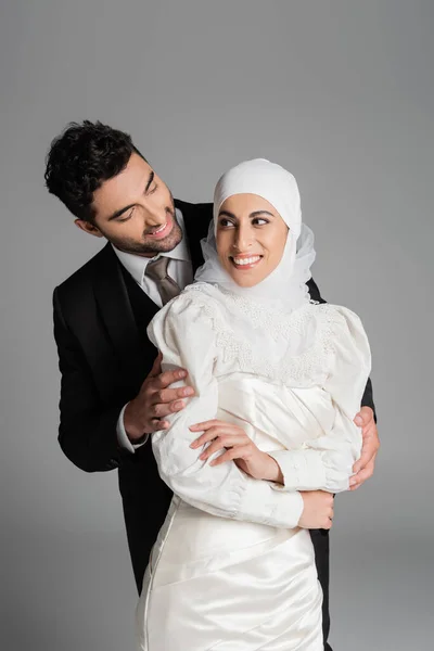 Felice sposo in piedi dietro sposa allegra in abito da sposa isolato su grigio — Foto stock