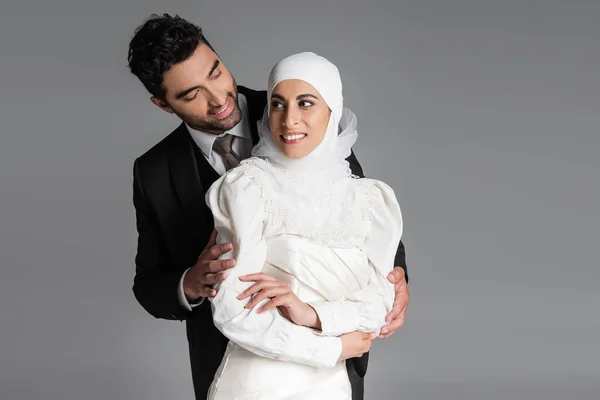Sorridente sposo in piedi dietro sposa allegra in abito da sposa isolato su grigio — Foto stock