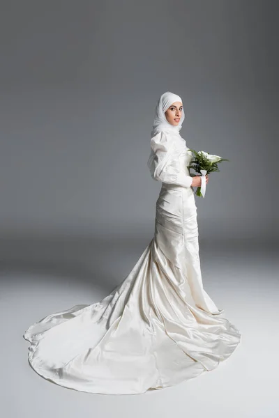 Vue pleine longueur de mariée musulmane dans une robe de mariée magnifique debout avec bouquet sur gris — Photo de stock