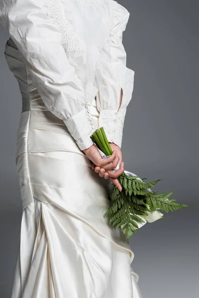 Cropped view of bride in gorgeous wedding dress standing with bouquet behind back isolated on grey — Stock Photo