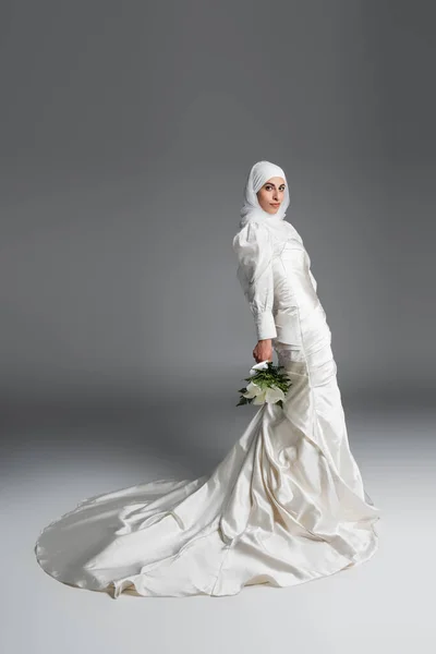 Полный вид мусульманки в великолепном свадебном платье, стоящей с букетом на сером — стоковое фото