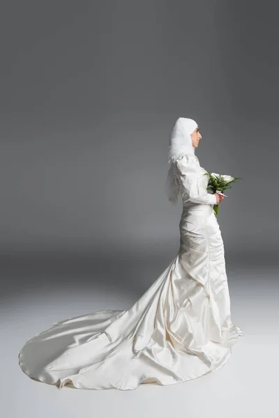 Vista lateral de la mujer musulmana feliz en vestido de novia magnífico de pie con ramo en gris - foto de stock