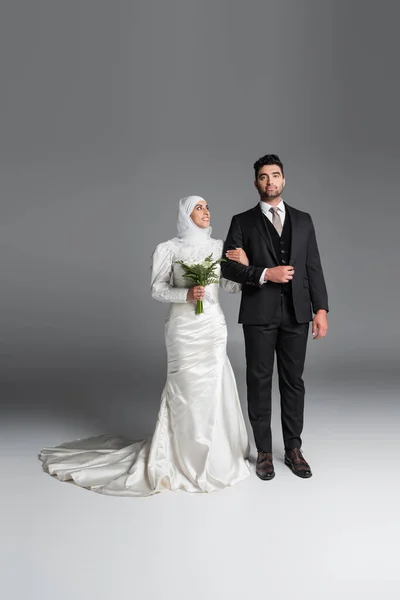 Longitud completa del novio en traje de pie con la novia musulmana feliz con ramo de flores de lirio de la cala en gris - foto de stock