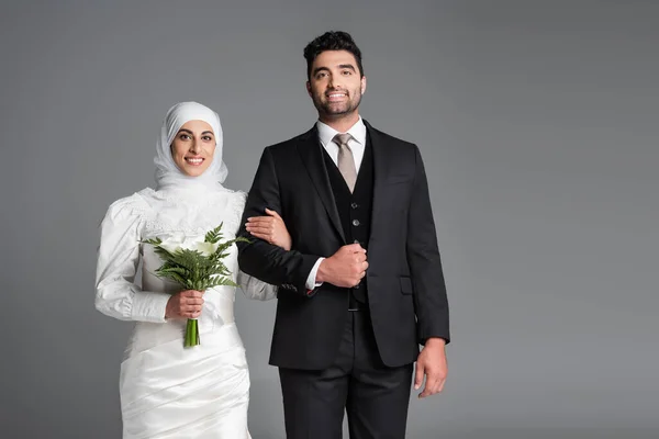 Портрет веселого жениха в костюме стоящей мусульманской невесты со свадебным букетом цветов калла лилии, изолированных на сером — стоковое фото