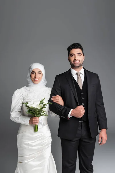Портрет счастливого жениха в костюме стоящей мусульманской невесты со свадебным букетом цветов калла лилии, изолированных на сером — стоковое фото