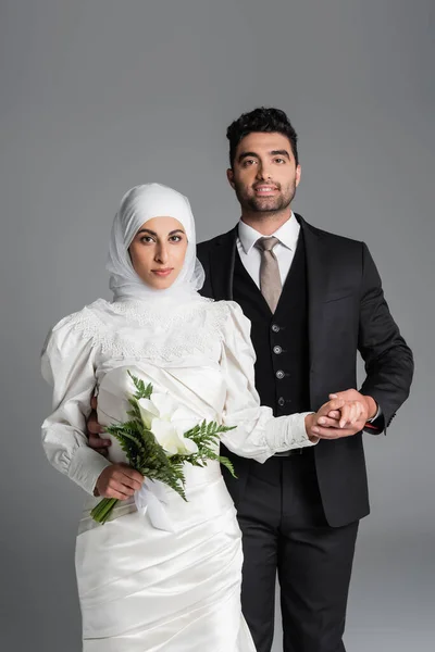 Happy groom in suit holding hand of muslim bride with wedding bouquet of calla lily flowers isolated on grey — Stock Photo