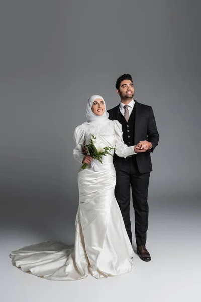 Повна довжина щасливого нареченого в костюмі тримає руку мусульманської нареченої з весільним букетом з каллі квіти лілії на сірому — стокове фото