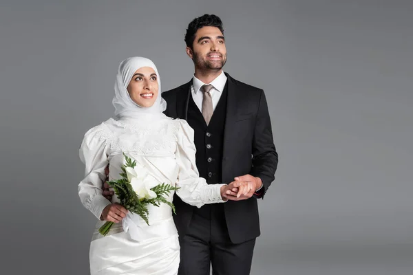 Happy groom in suit holding hand of dreamy muslim bride with wedding bouquet of calla lily flowers isolated on grey — Stock Photo