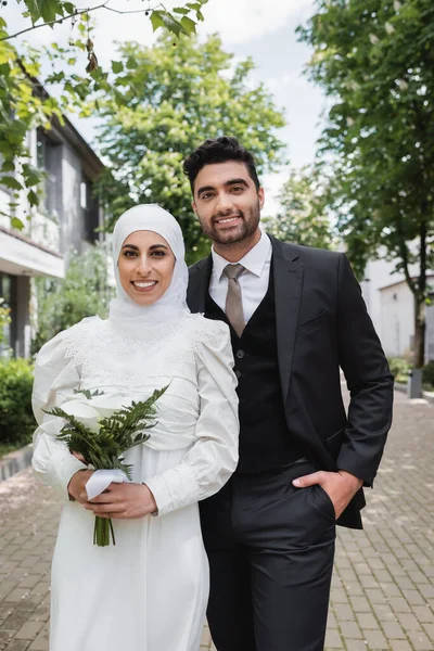 Noivo feliz posando com a mão no bolso perto de noiva muçulmana no hijab com buquê de casamento — Fotografia de Stock