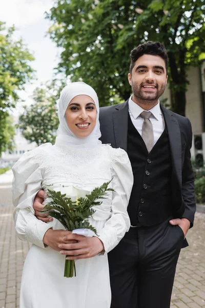 Novio alegre posando con la mano en el bolsillo cerca de la novia musulmana en hijab con ramo de boda - foto de stock