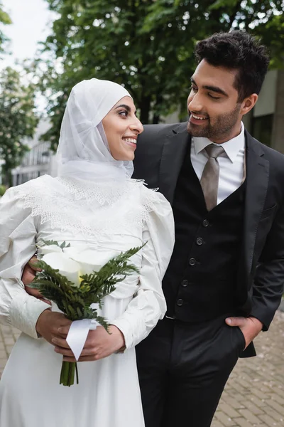 Novio feliz posando con la mano en el bolsillo y mirando a la novia musulmana en hijab con ramo de bodas - foto de stock