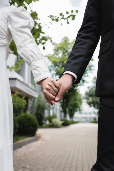 Cropped view of newlyweds in wedding dress and formal wear holding hands outdoors — Stock Photo