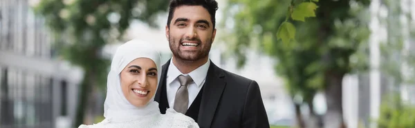 Marié heureux étreignant mariée musulmane dans le hijab souriant et regardant la caméra, bannière — Photo de stock