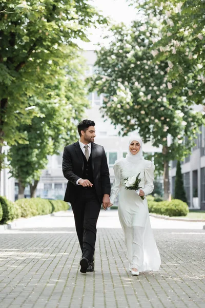 Novio feliz cogido de la mano con la novia musulmana con ramo de bodas y caminar juntos - foto de stock