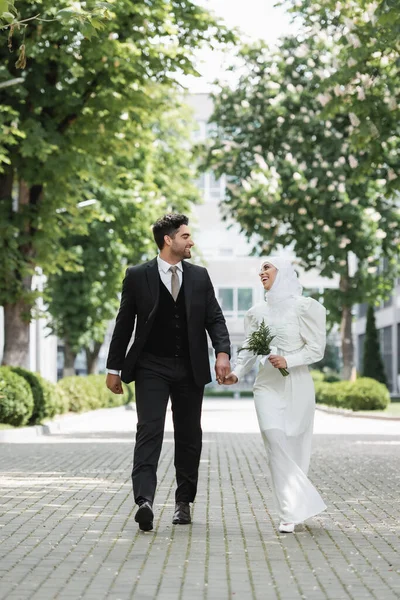 Joyful groom holding hands with muslim bride with wedding bouquet and walking together — Stock Photo