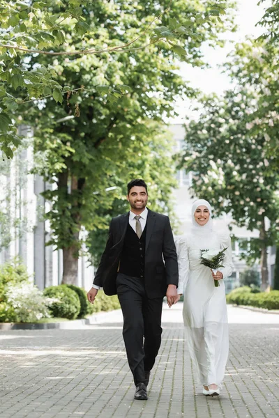 Marié heureux tenant la main avec la mariée musulmane souriante avec bouquet de mariage et marchant ensemble — Photo de stock